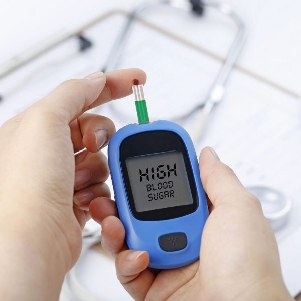Hand holding a blood glucose meter measuring blood sugar, the background is a stethoscope and chart file